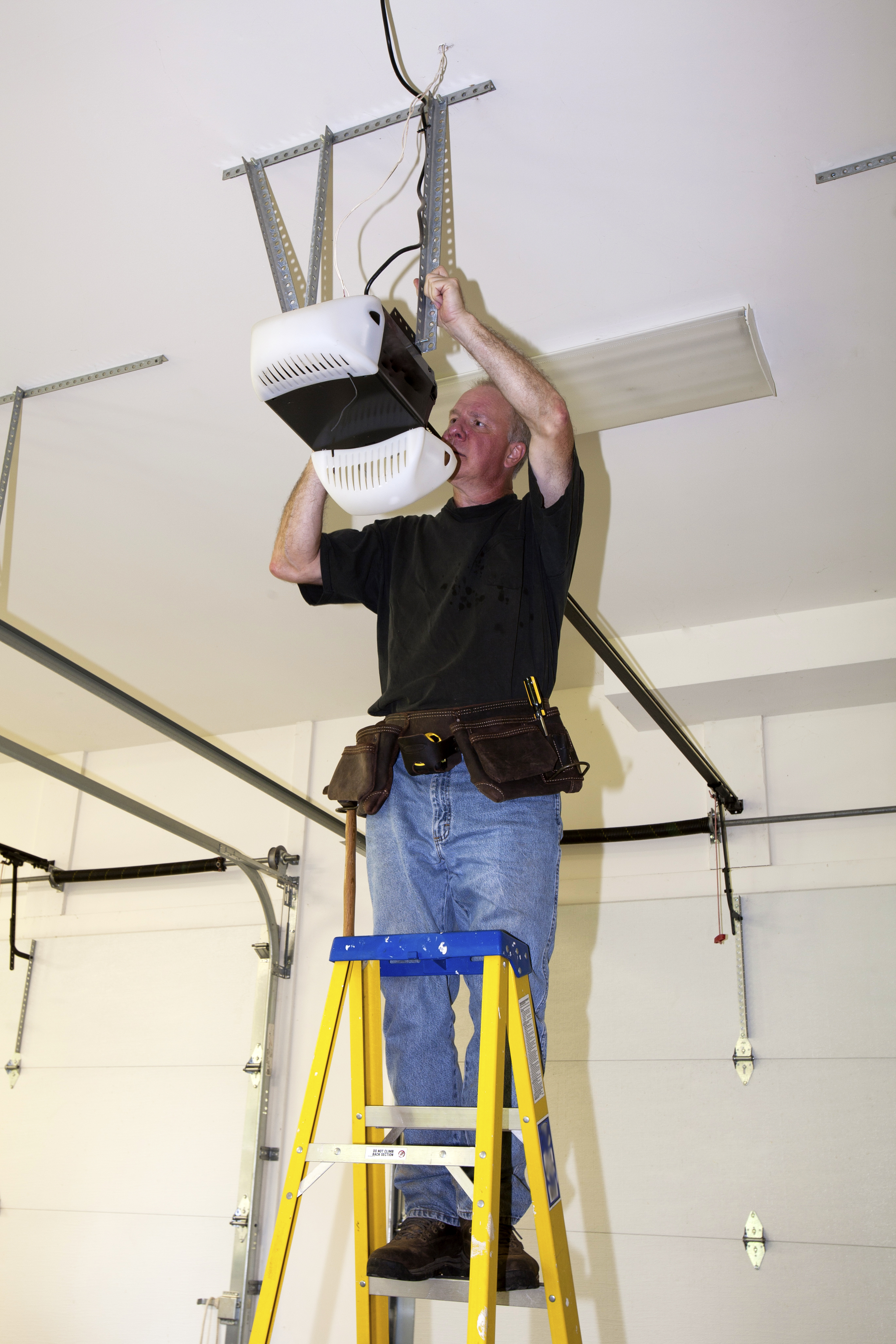 Contractor-Repairing-Broken-Garage-Door-Opener Man standing on a ladder fixing a mechanical garage door opener